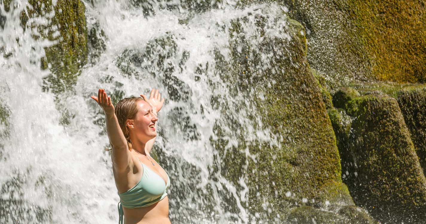 A student stretches their arms to the sky before a waterfall on Opal Creek