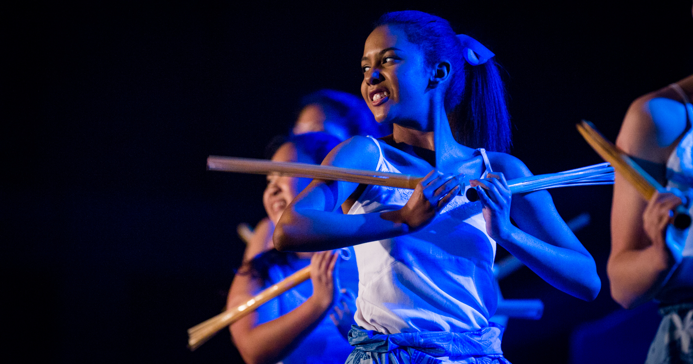 In blue light, a woman smiles while holding drum-stick like objects while dancing in the luau