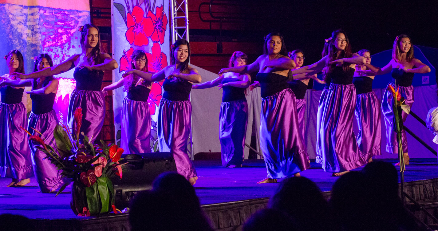 two lines of women each with their hands extended to their right dance onstage at the luau