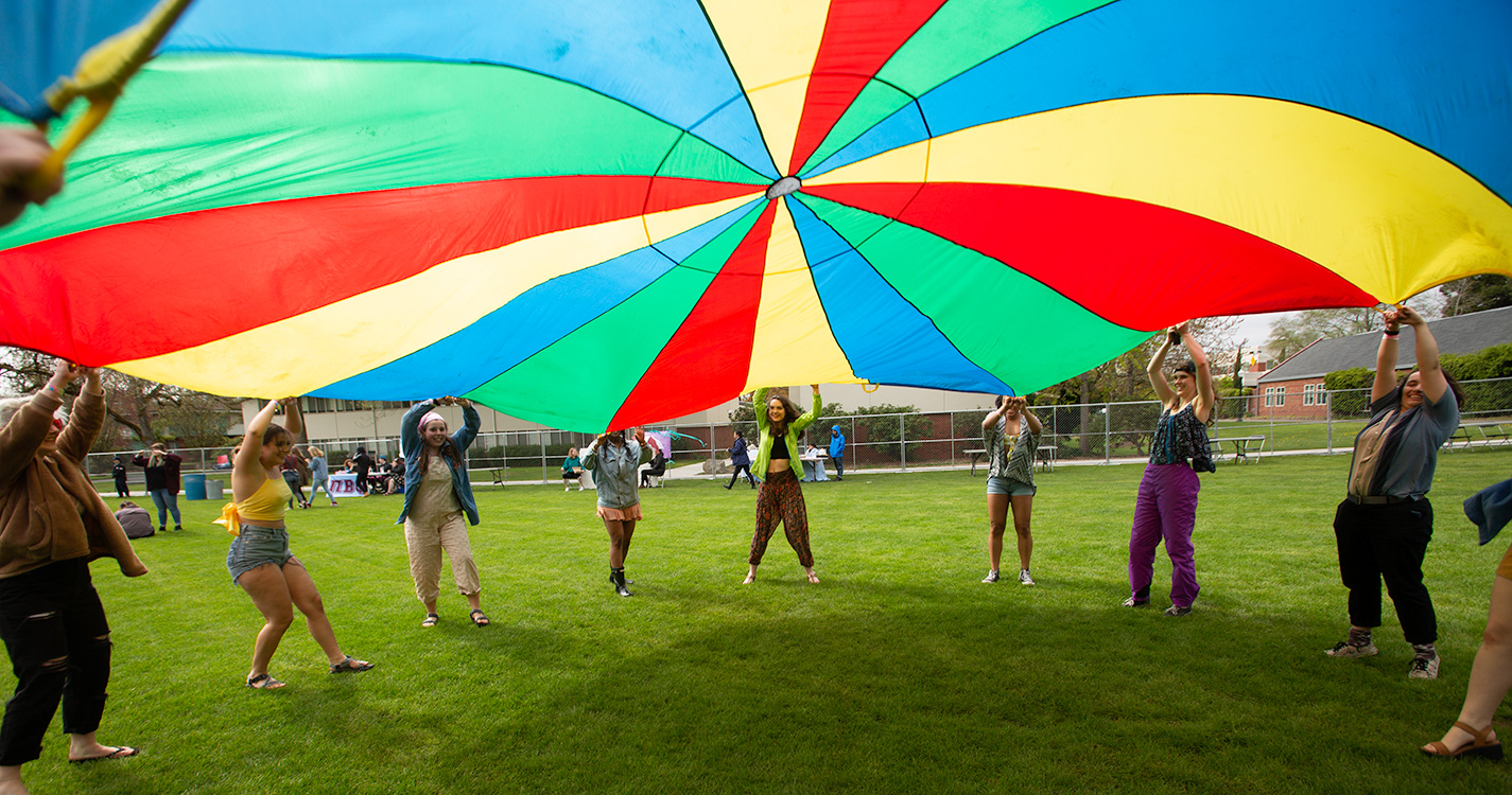 Students standing in a circle raise a rainbow colored parachute to the sky.
