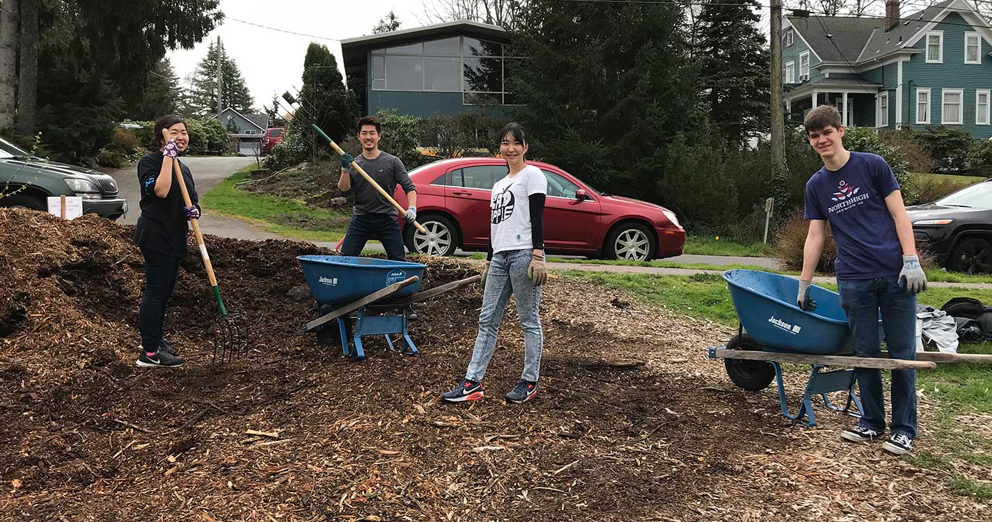 Students work in a Seattle park moving mulch with shovels and a wheel barrow.