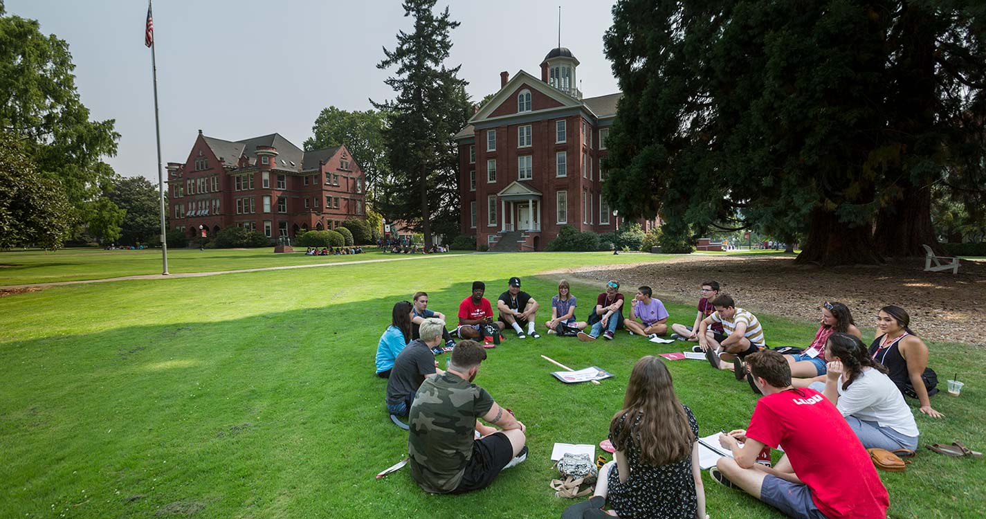 A small group of new students sits in a circle in the shade of the Star Trees with Waller Hall in the background. 