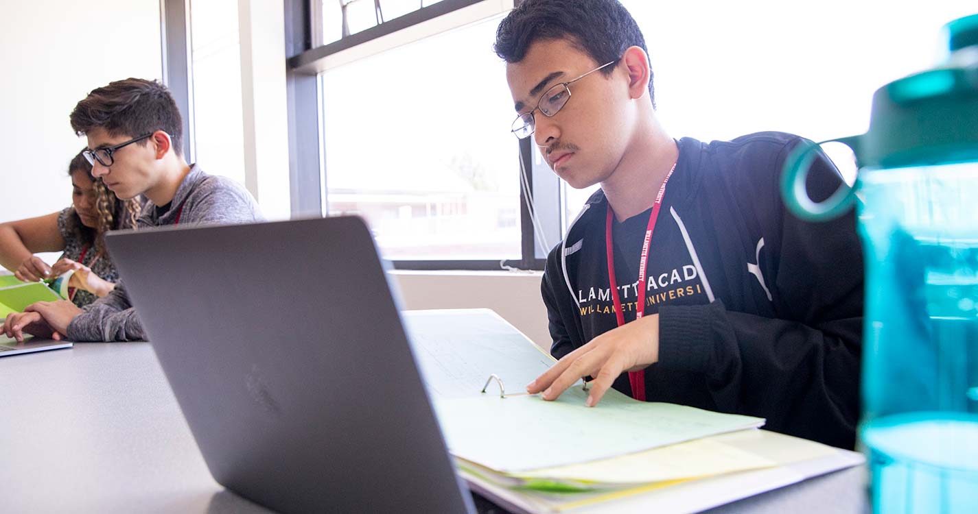 A student with an open laptop reads a notebook