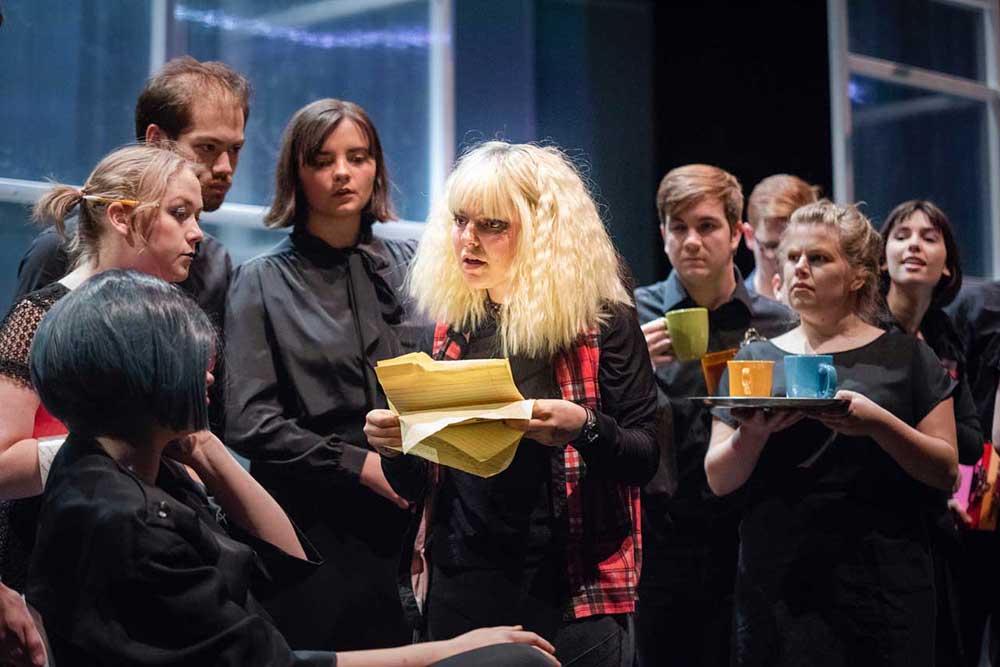 A woman with blonde hair and dark eye makeup stands holding pages ripped from a yellow pad, she's surrounded by eight men and women in black a few are holding coffee cups