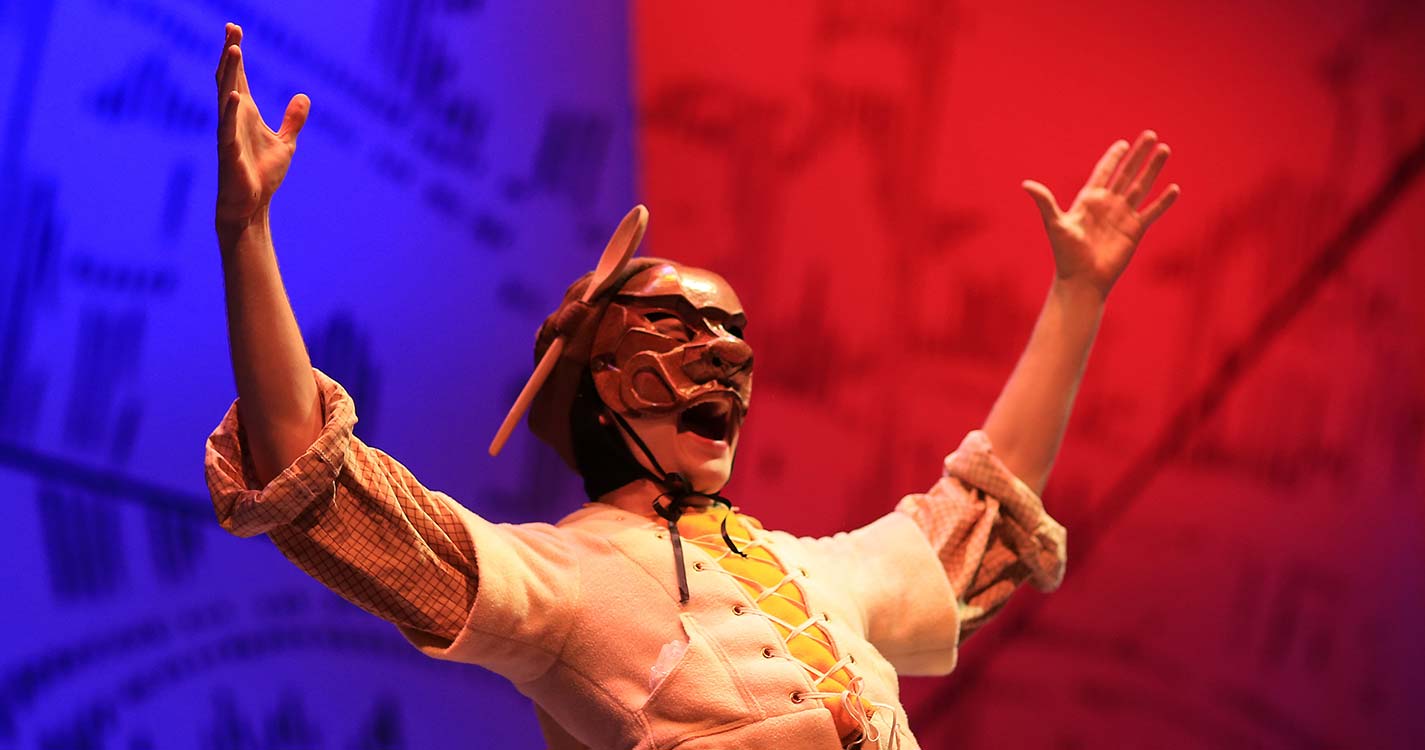 Character wearing wooden mask with hands outstretched in a Y before a blue and red backdrop