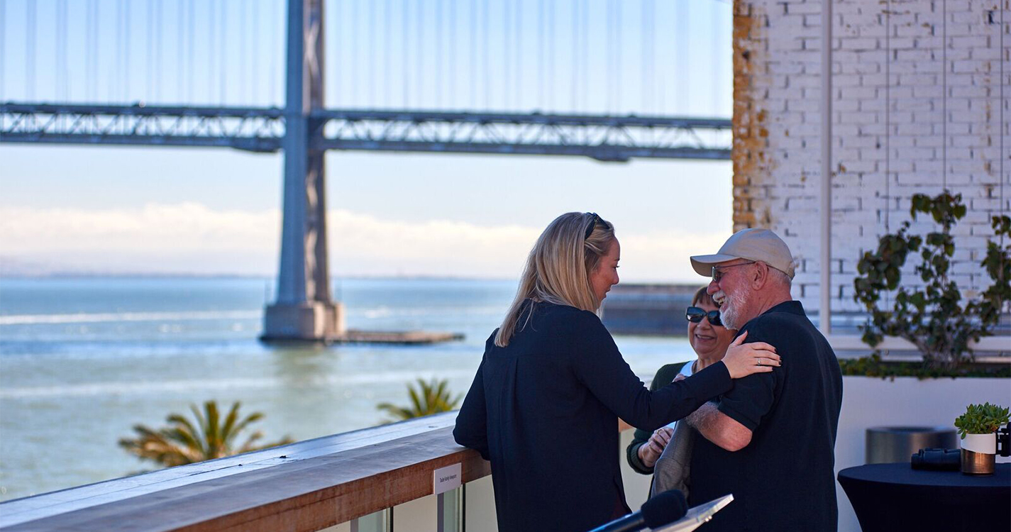Kate Steffy speaks with two people on a patio with a waterway and suspension bridge in the background.