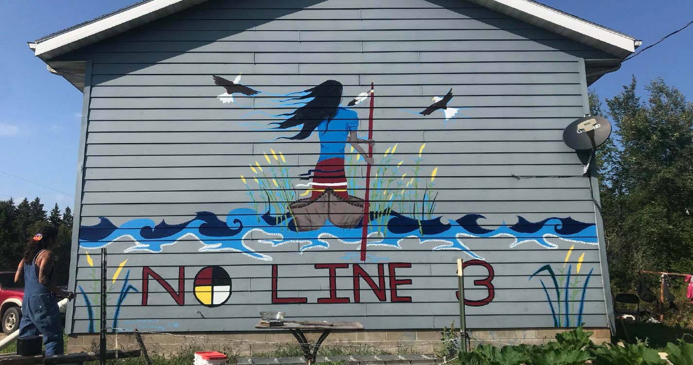 Painting on the side of a house of person with long black hair standing on a canoe surrounded by willows and water with two bald eagles overhead with phrase, "No Line 3."