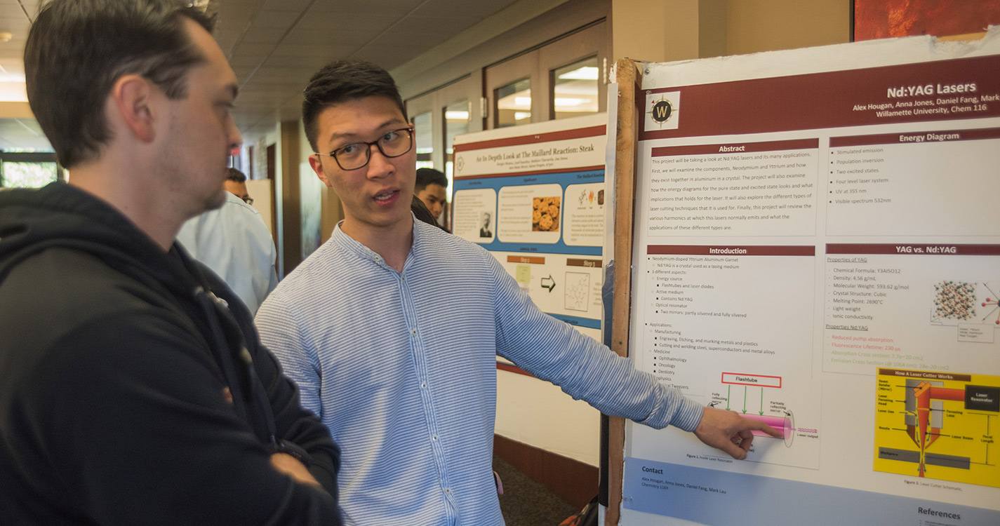 Student points at research on a poster while speaking to another