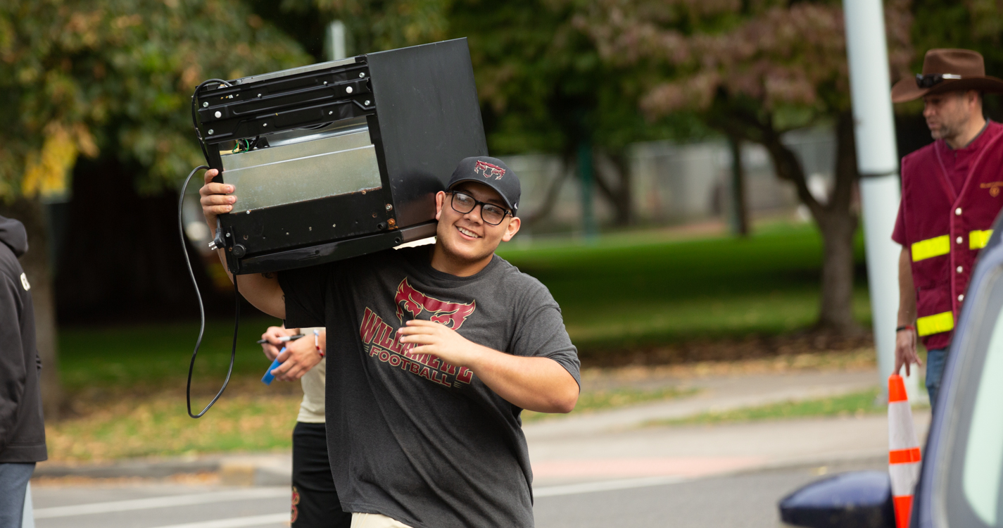 A student wearing a Bearcats shirt carries a refrigerator on their shoulder