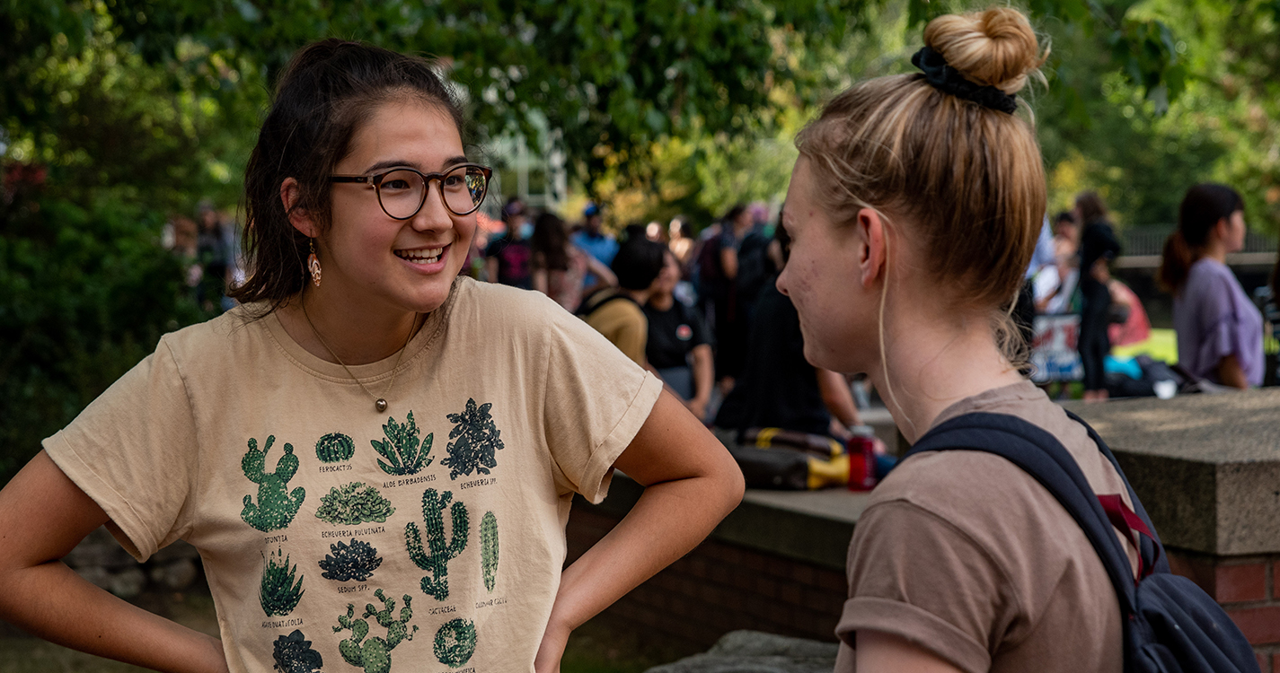 A student wearing a shirt with numerous differently shaped cacti talks to another student