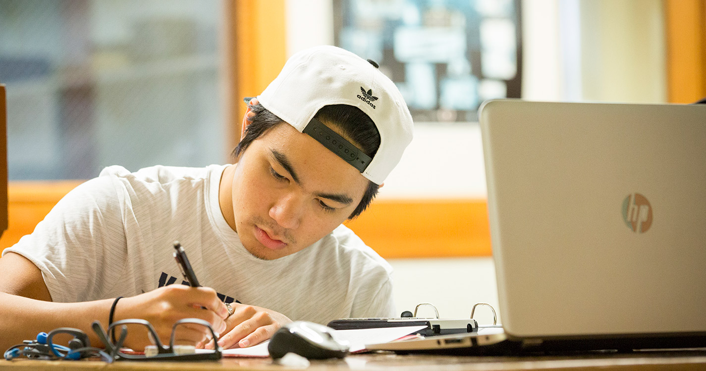 Student writes on a piece of paper with an open laptop in the foreground