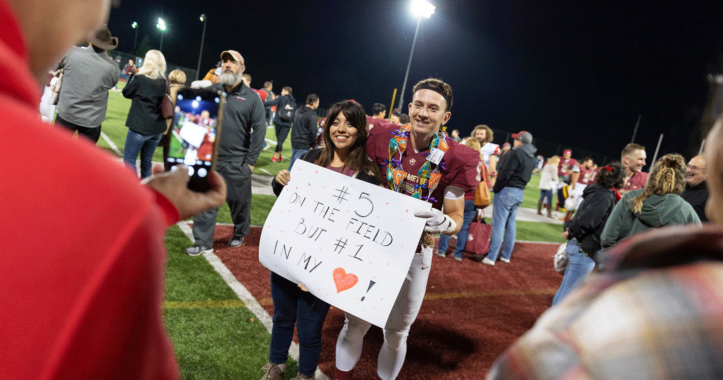 A football player stands next to a person holding a sign "#5 on the field but #1 in my heart!"