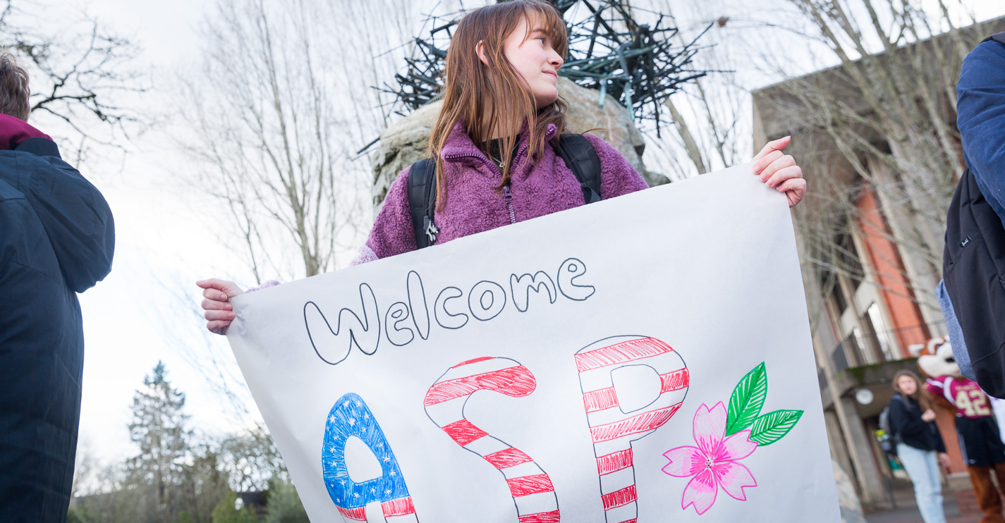 A student holds a sign that says Welcome ASP in red, white and blue with a pink cherry blossom
