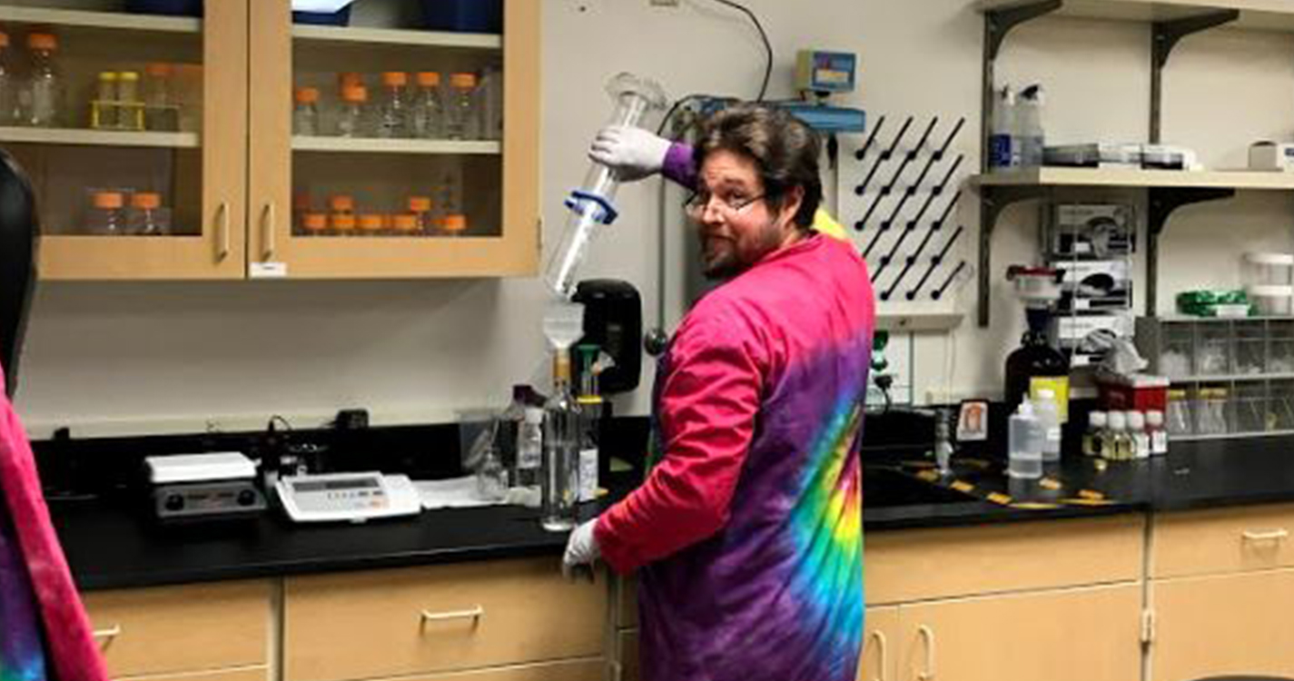 Lab worker in tie-dye lab coat pours solution from a beaker