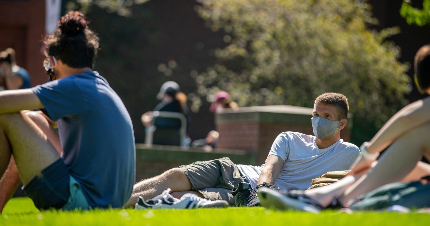 Students in facemasks lounging on the lawn in front of Mill Stream