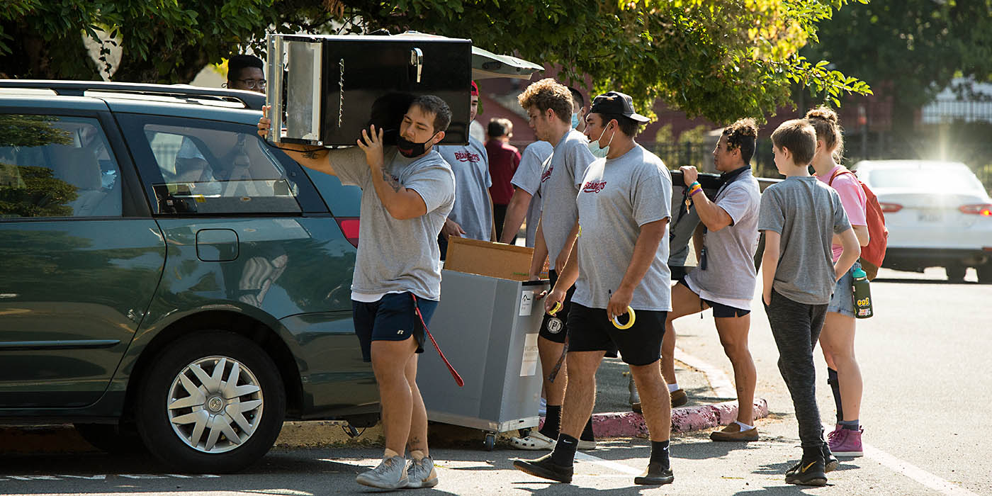 Some student athletes haul a mini-fridge into the dorms for a new student in front of Baxter Hall.