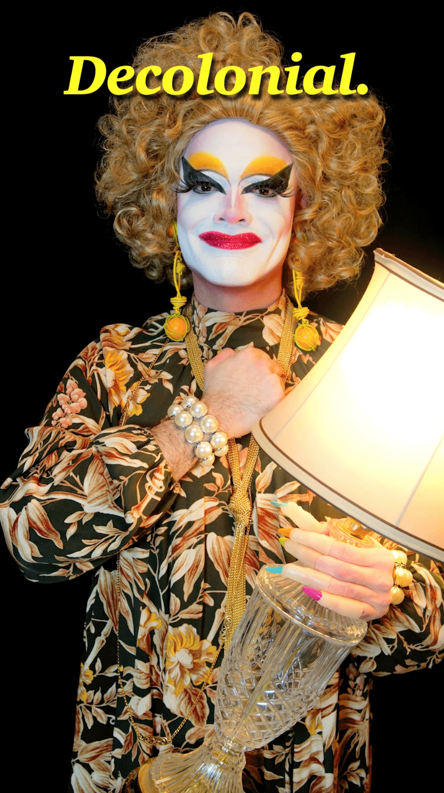 A drag queen clown with a large blonde wig holds a lamp 